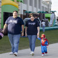 Trey and Josie Clark walk beside their young son, Andy, who wears a Spider-Man costume and holds Josie’s hand. Bounce houses and other costumed attendees are behind them at Fletcher’s Fall Fest.