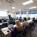 Attendees seated at classroom tables listen as Ron C. Henderson presents beside a projected slide at Fletcher Technical Community College’s Bayou Region Continuing Education Symposium.