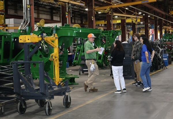 H. L. Bourgeois High School students gather around a tour guide beside green manufacturing equipment on the John Deere factory floor in Thibodaux.