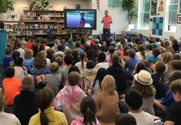 Man standing in front of a library full of seated students talking in front of a digital display of a small boy.