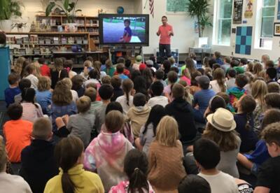 Man standing in front of a library full of seated students talking in front of a digital display of a small boy.