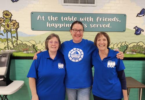 Three ladies wearing blue shirts with their arms around one another.