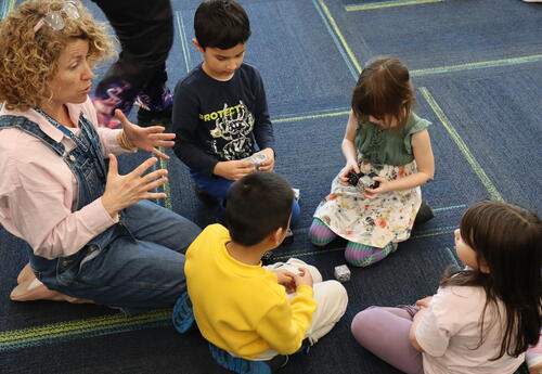 Students and a teacher sitting on a blue rug building a cube shaped robot