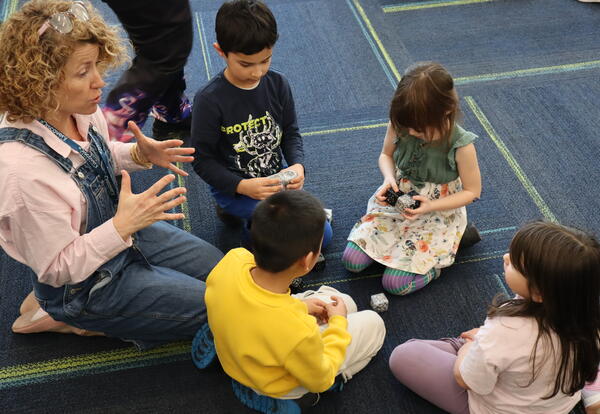 Students and a teacher sitting on a blue rug building a cube shaped robot