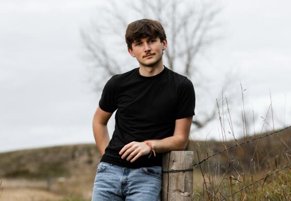 Everett “Skippy” McRae-Lorenz in a black shirt and jeans leaning against a wooden post in a grassy field.