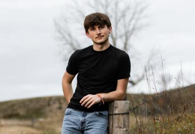 Everett “Skippy” McRae-Lorenz in a black shirt and jeans leaning against a wooden post in a grassy field.