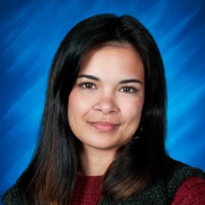 Professional headshot of a woman with dark hair, wearing a burgundy sweater and black textured vest on a blue background.