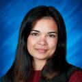 Professional headshot of a woman with dark hair, wearing a burgundy sweater and black textured vest on a blue background.