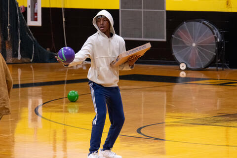 An eighth-grade student holds a dodgeball and a pizza box during a game. 