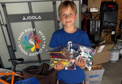 A child in a blue shirt stands indoors holding bags filled with colored pencils and dry erase markers.