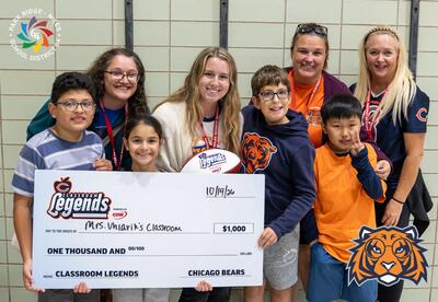 Teacher holding a Chicago Bears branded football with Students surrounding her holding a Check on behalf of the Chicago Bears to Utilize towards Uhlarik's classroom.