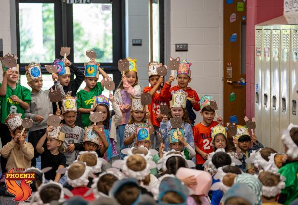Students performing a nursery rhyme show. The students are wearing hats that they made, with a drawing of a vibrant clock. They are also holding up a craft that is a brown cat on a popsicle stick. Their peers are sitting watching them while they are weari